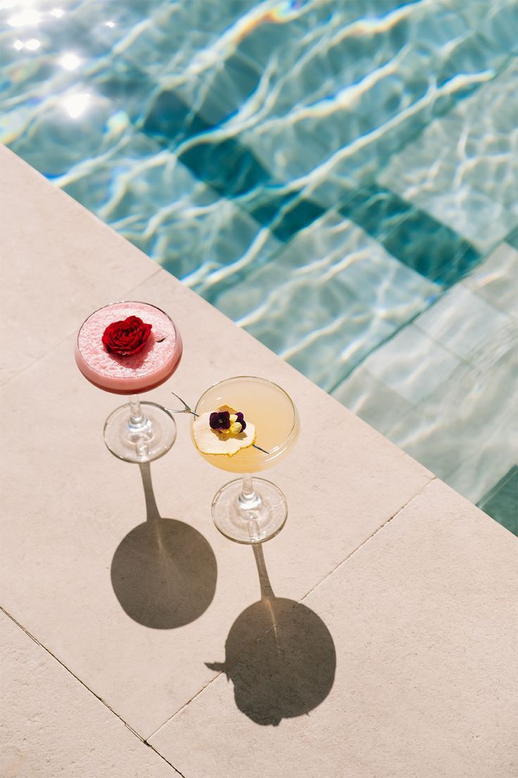 two wine glasses sitting next to each other near a swimming pool