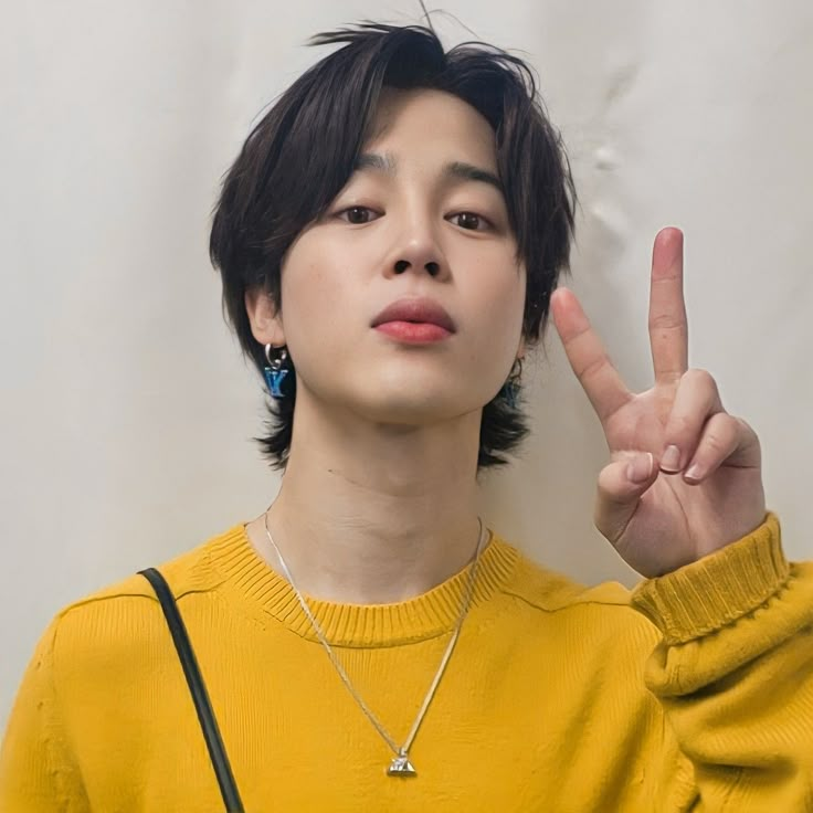 a young man making the peace sign with his hand while wearing a yellow sweater and black cross - body bag