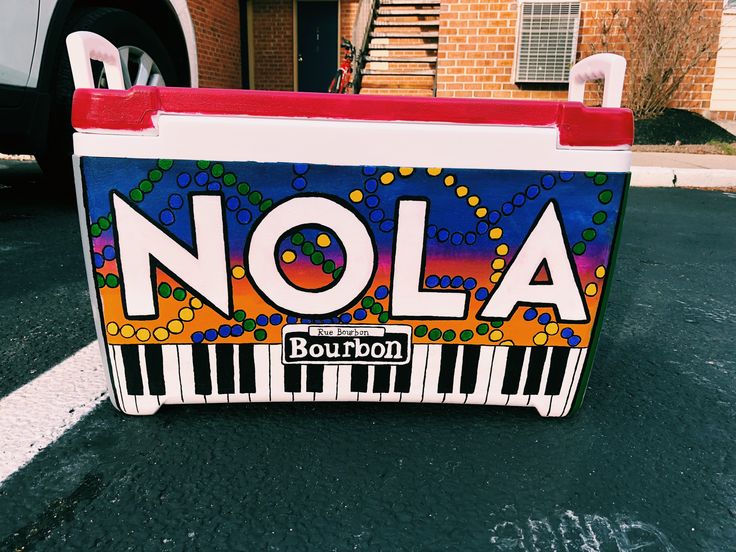 a piano painted with the word nola on it in front of a building and parked car