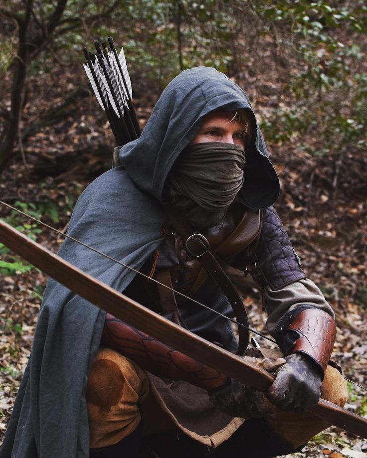 a man dressed in medieval clothing holding a bow and arrow while crouching on the ground