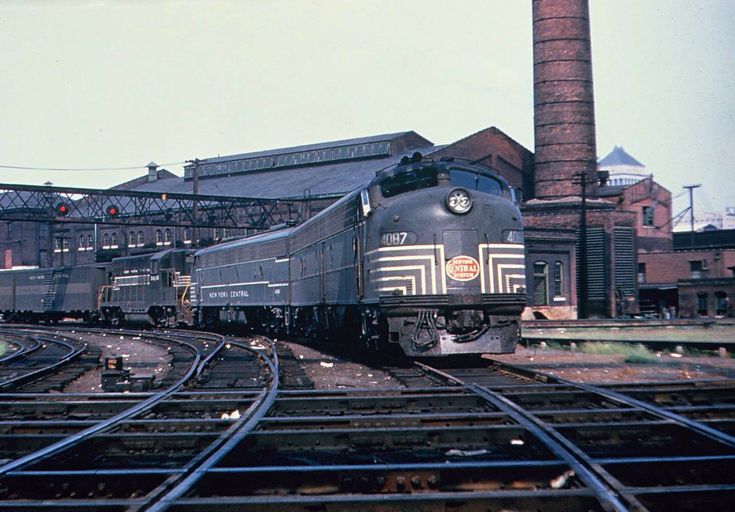 the train is going down the tracks near an industrial building and smokestack tower