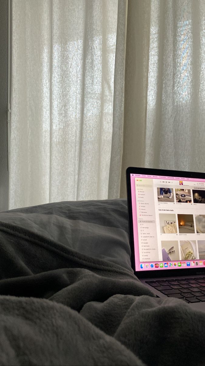 a laptop computer sitting on top of a bed next to a window with white curtains