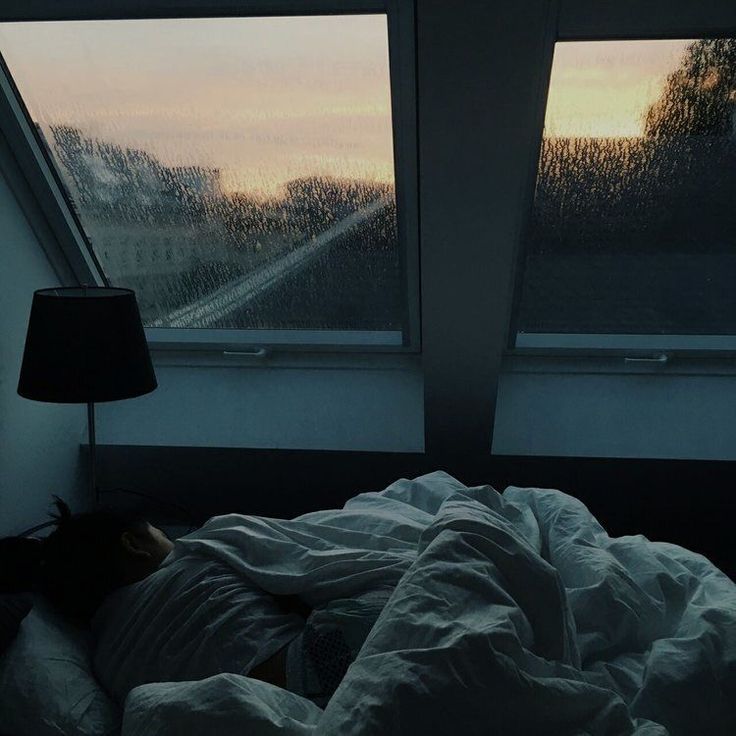 a person laying in bed under a blanket next to two windows with rain falling on them