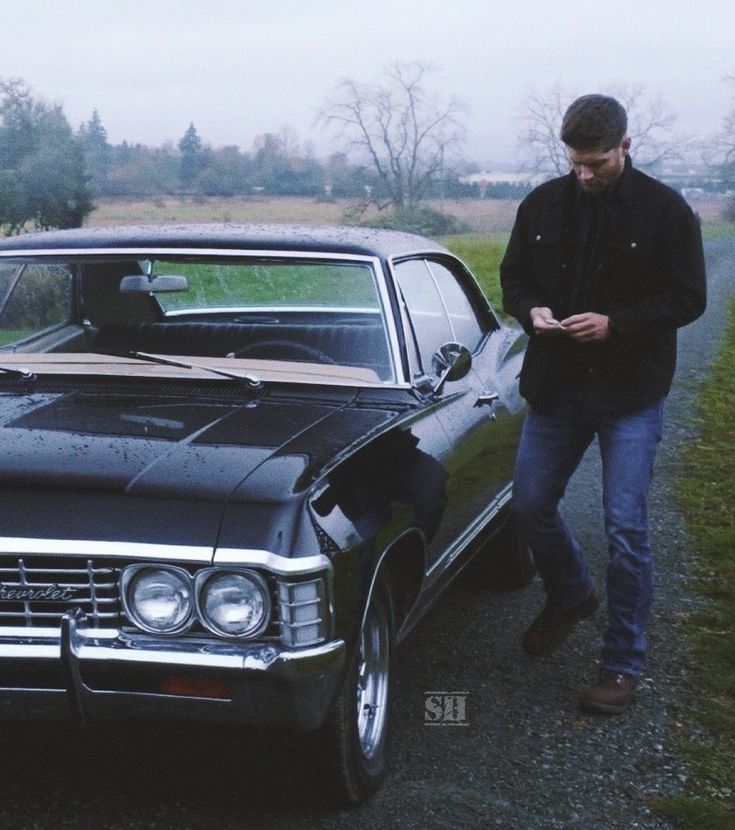 a man looking at his cell phone next to an old car on the side of the road