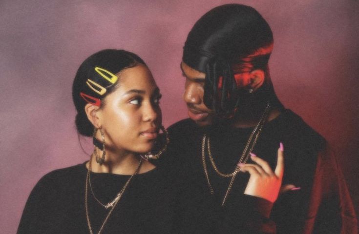 a man and woman standing next to each other in front of a purple background wearing black clothing