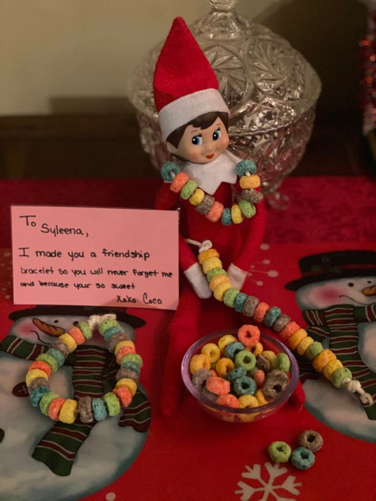 an elf is sitting on the table with some cereal beads and a sign that says it's christmas