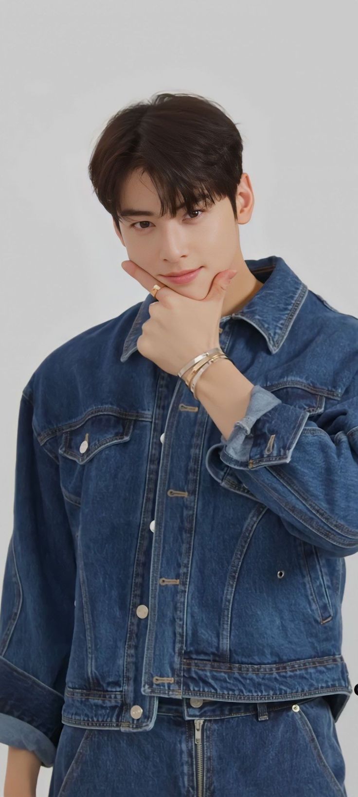 a young man wearing a denim jacket and jeans with his hand on his chin, posing for the camera