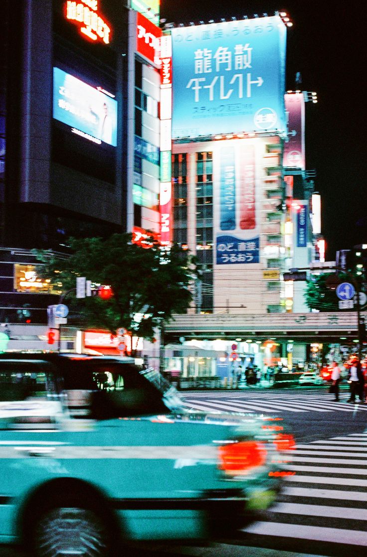 a green car driving down a street next to tall buildings with neon signs on them