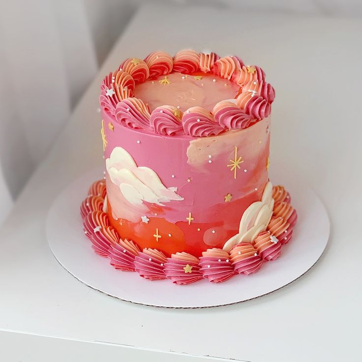 a pink and red cake with clouds on it sitting on a white plate next to a box