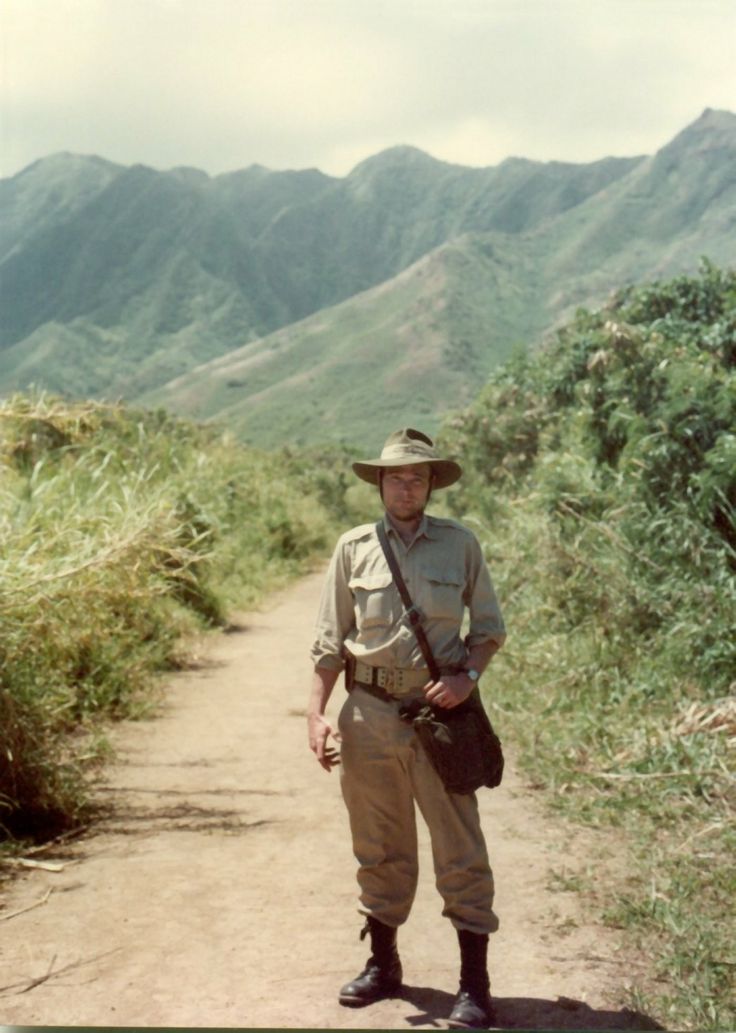 a man in uniform standing on a dirt road with mountains in the backgroud