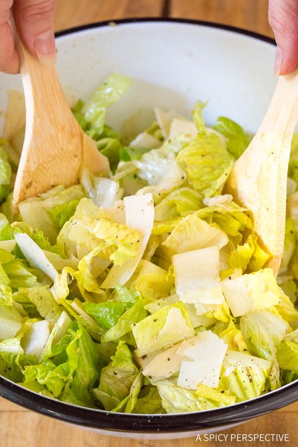 two hands are holding wooden spoons over a bowl of lettuce and cheese