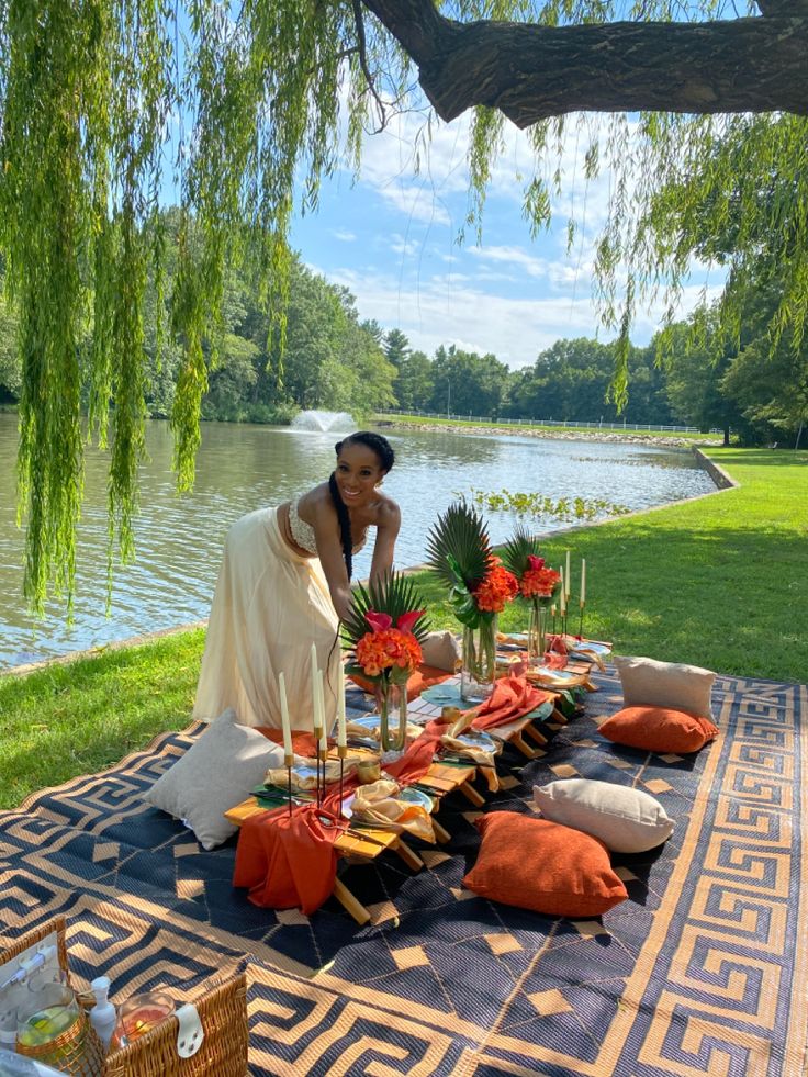 a woman is setting up an outdoor table with flowers and candles on it in front of a lake