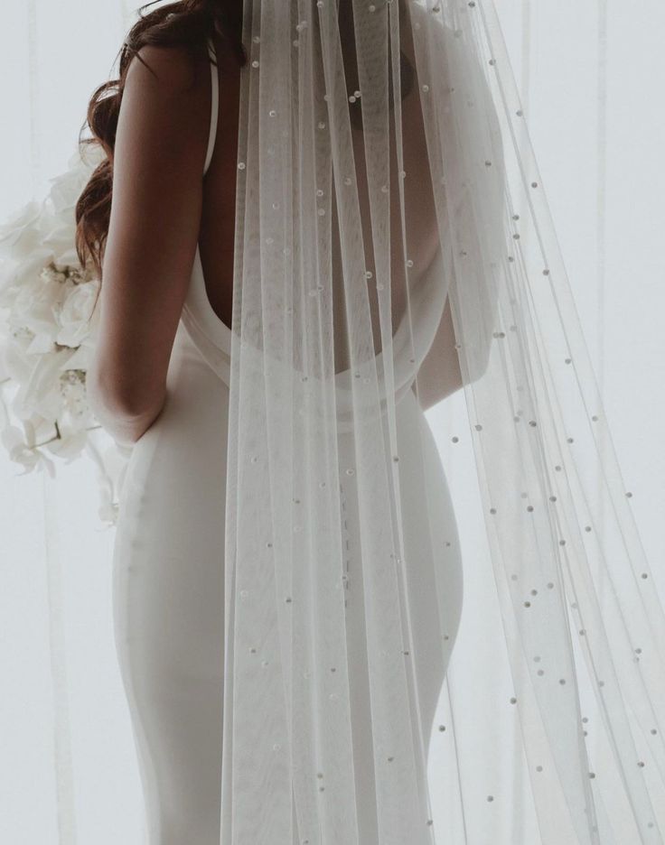 a woman in a white wedding dress holding a bouquet of flowers behind her back and veil over her head