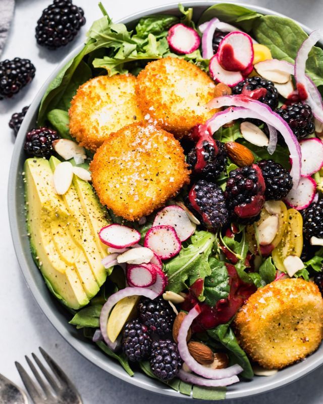 a salad with blackberries, avocado and croutons in it on a white table