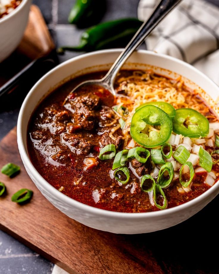 a bowl of chili soup on a cutting board with a spoon in it and green peppers