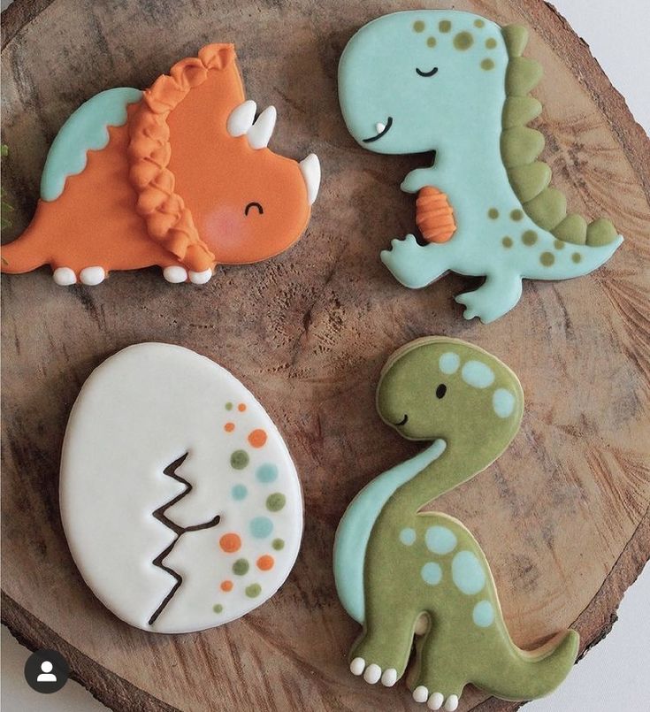 three decorated cookies sitting on top of a wooden plate next to an egg and dinosaur