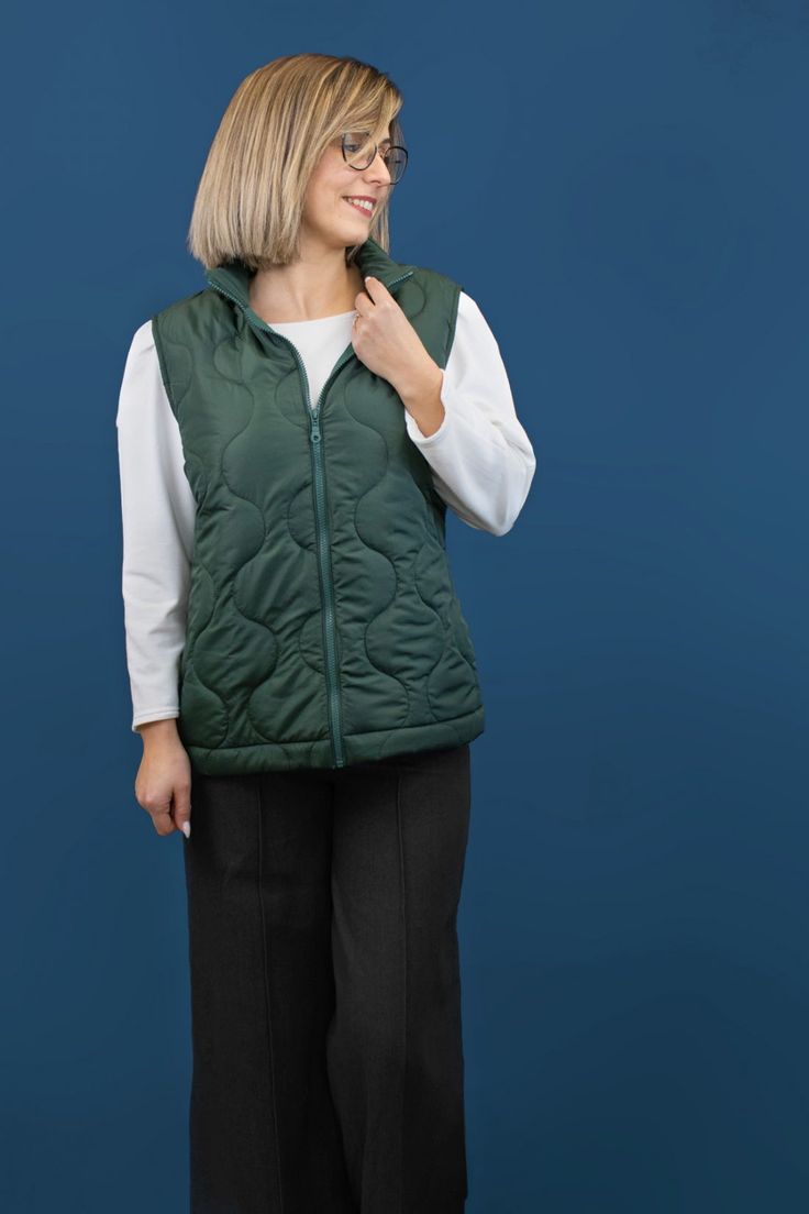 a woman standing in front of a blue background wearing a green vest and black pants