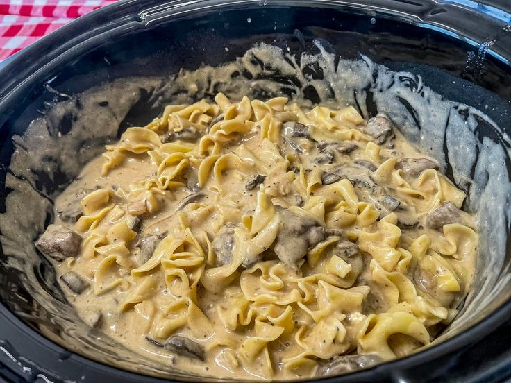 a crock pot filled with pasta and meat in gravy on top of a checkered table cloth