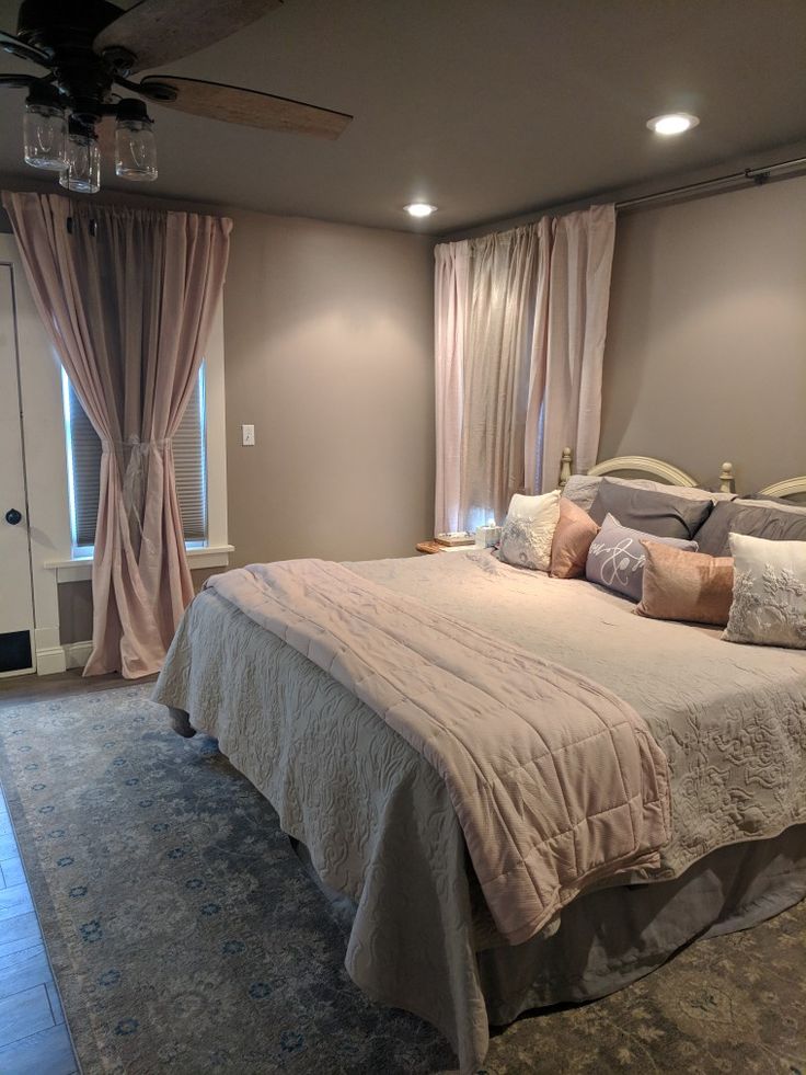 a bedroom with a bed, ceiling fan and two windows in the room that have curtains on them