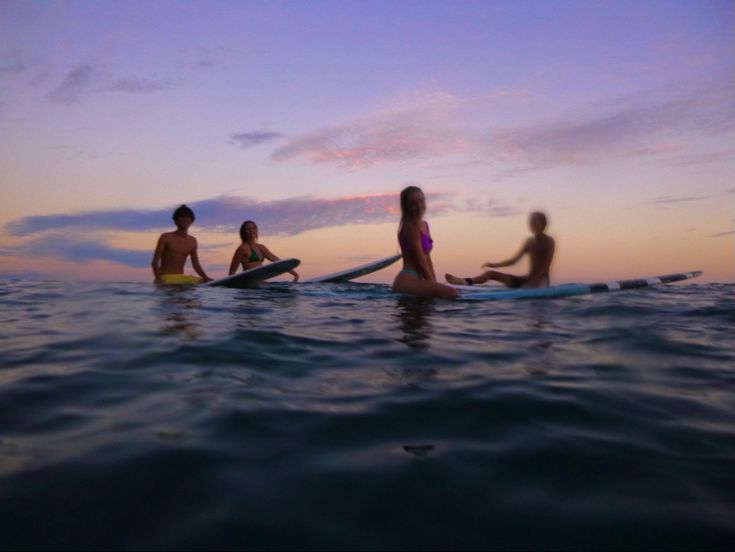 three people are sitting on surfboards in the water at sunset or dawn, with one person standing up