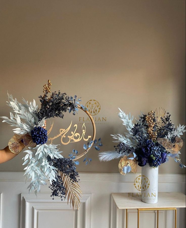 two vases with blue flowers and greenery in them on a table next to a wall