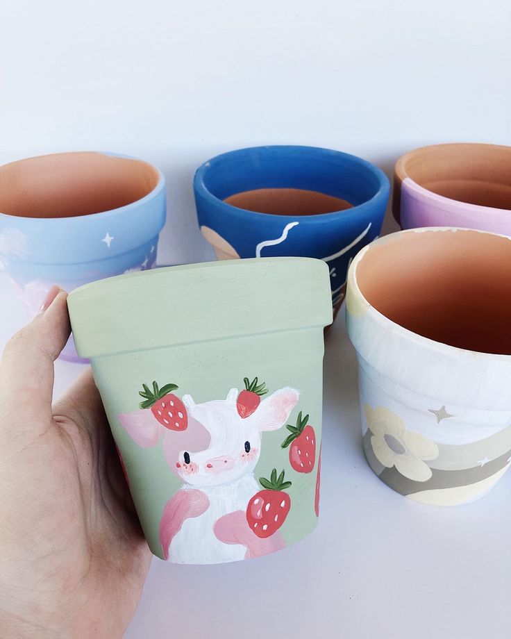 a hand holding an empty cup next to four different colored cups with strawberries on them