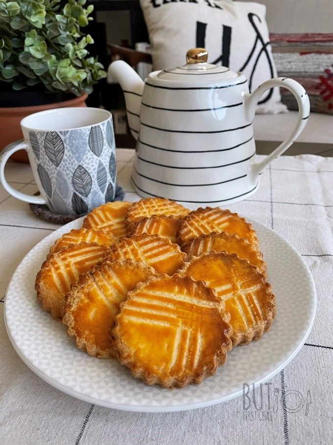 some cookies are sitting on a plate next to a cup and tea pot with a plant in the background