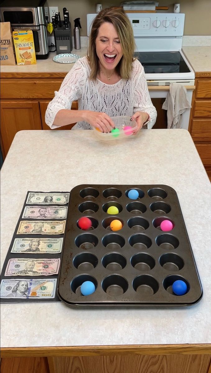 a woman sitting at a kitchen counter with a tray of cupcakes and dollar bills