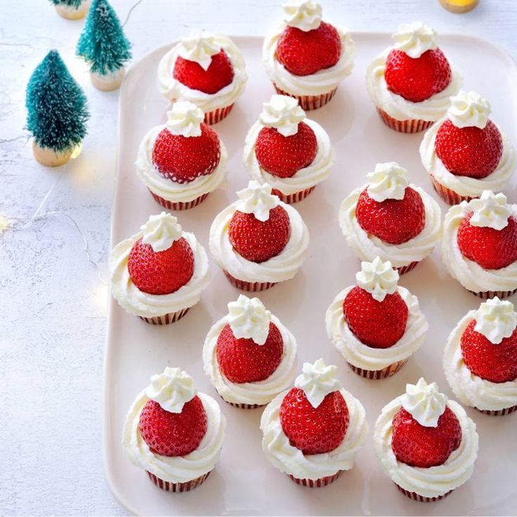 cupcakes with white frosting and strawberries arranged on a tray next to small christmas trees