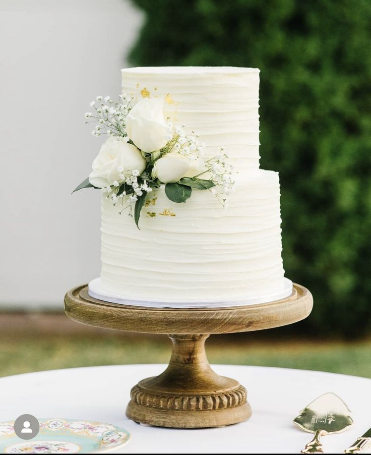 a wedding cake with white flowers and greenery sits on a wooden stand in front of a green bush