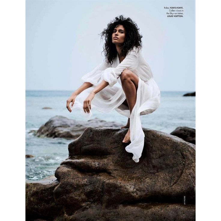 a woman sitting on top of a rock next to the ocean wearing a white dress