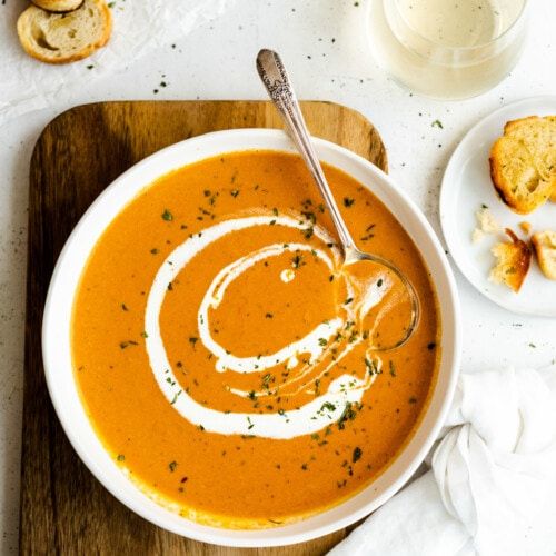 a bowl of carrot soup on a wooden cutting board with bread and wine in the background