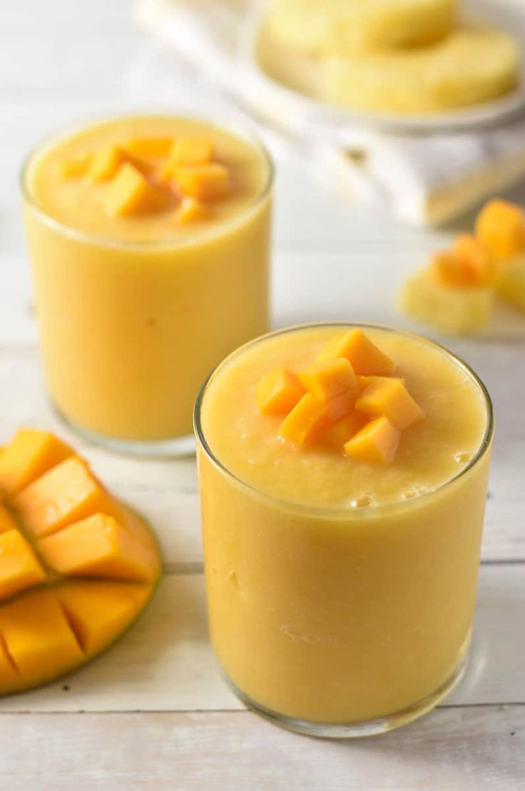 two glasses filled with mango smoothie on top of a white table next to sliced mangos