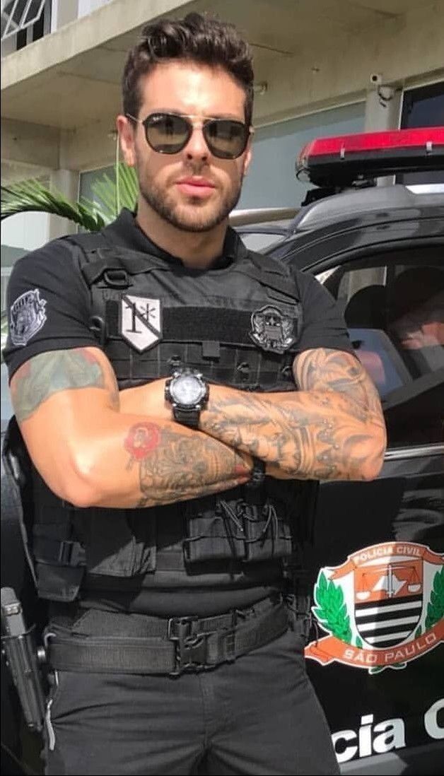 a man with his arms crossed standing in front of a police car and wearing sunglasses