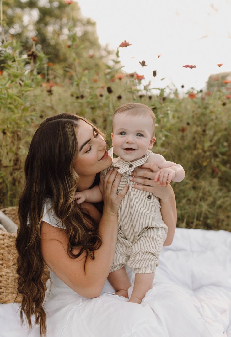 a woman holding a baby in her arms and kissing it's face with butterflies flying overhead