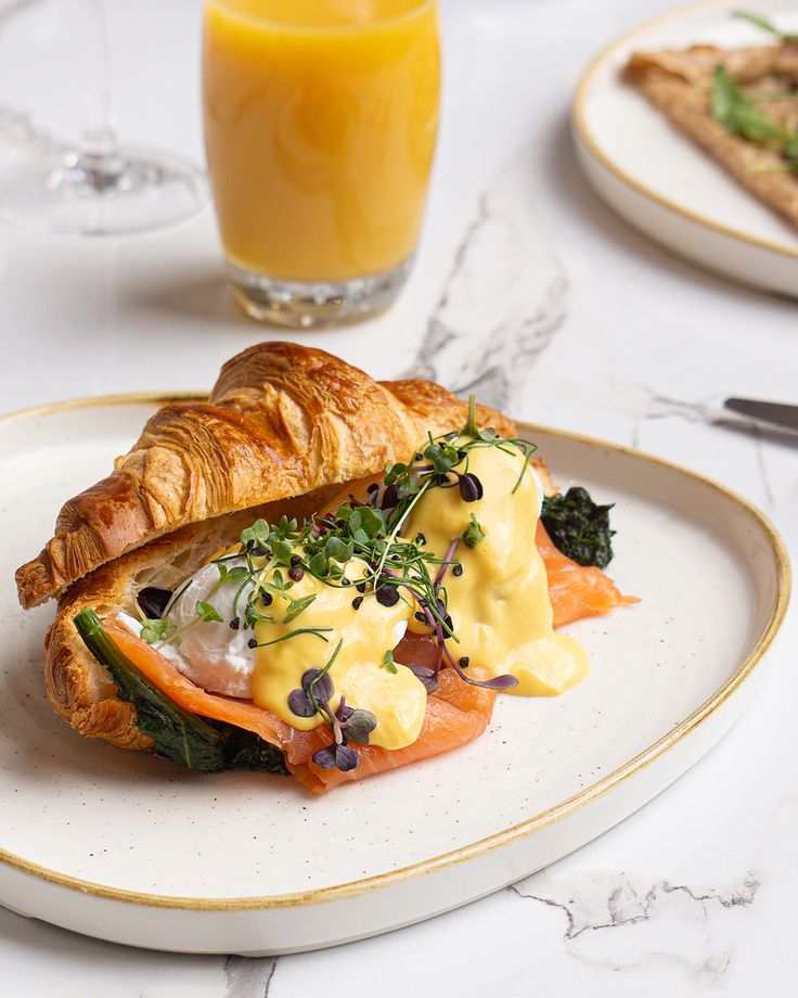 a white plate topped with a croissant covered in cheese and vegetables next to a glass of orange juice