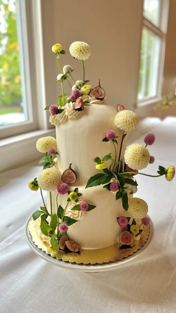 a three tiered white cake with flowers on the top and bottom is sitting on a table in front of a window