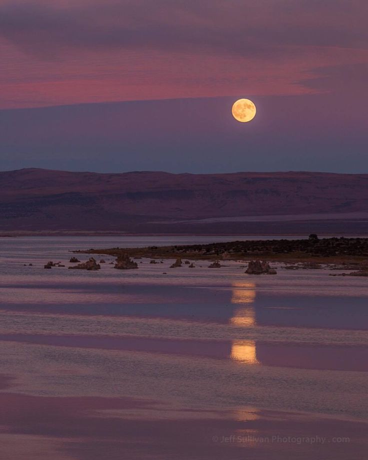 the full moon is setting over water with mountains in the background