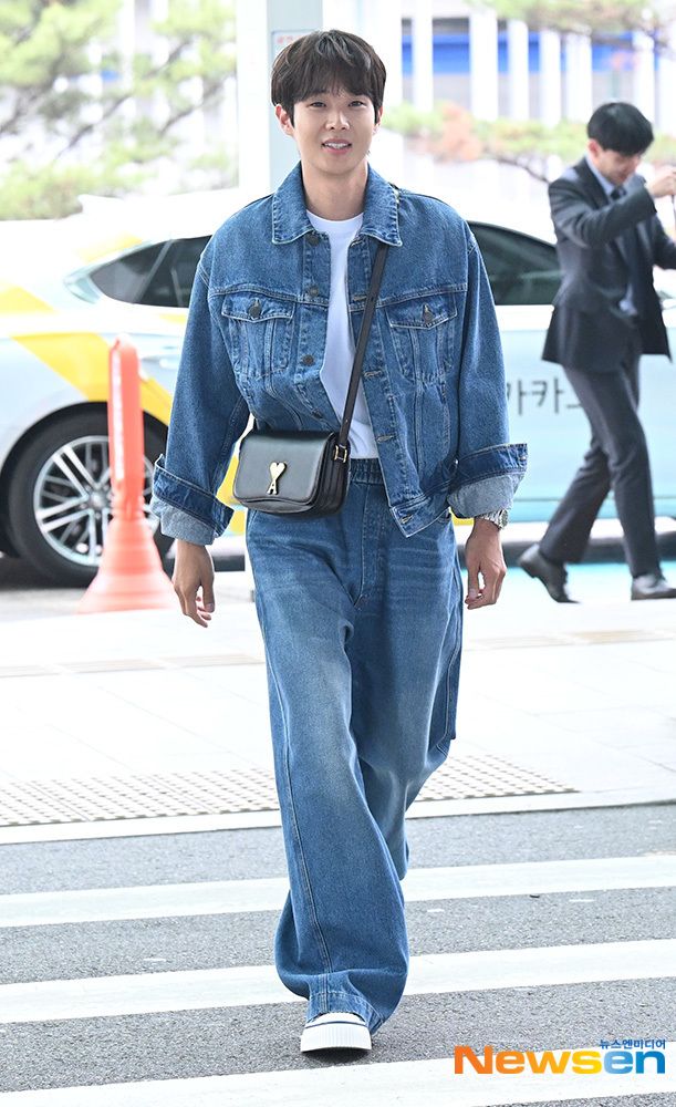 a man walking across a cross walk holding a cell phone in his hand and wearing a denim jacket