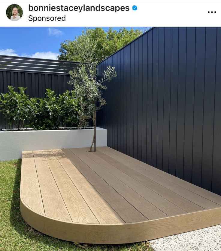 a wooden deck in front of a black wall and green grass with a small tree