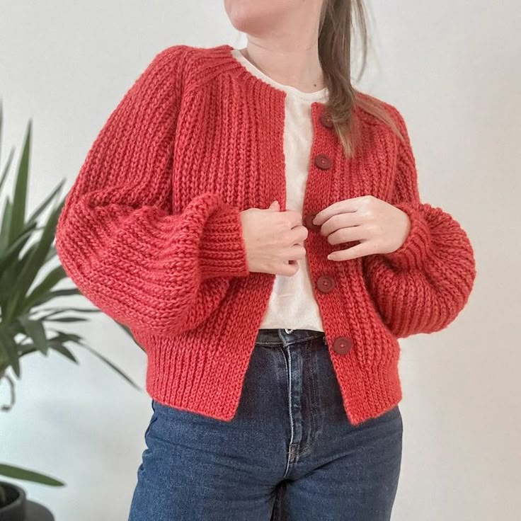 a woman standing in front of a plant wearing a red cardigan