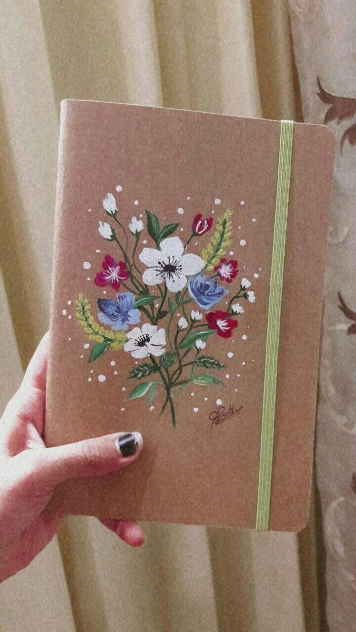 a hand holding a notebook with flowers painted on the front and side, in front of a curtain