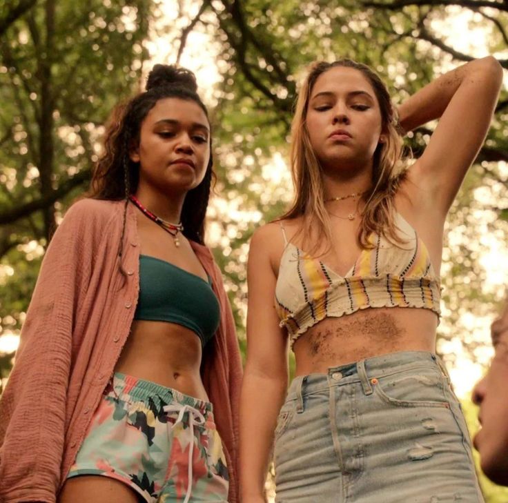 two women standing next to each other in front of trees and grass, one wearing a crop top