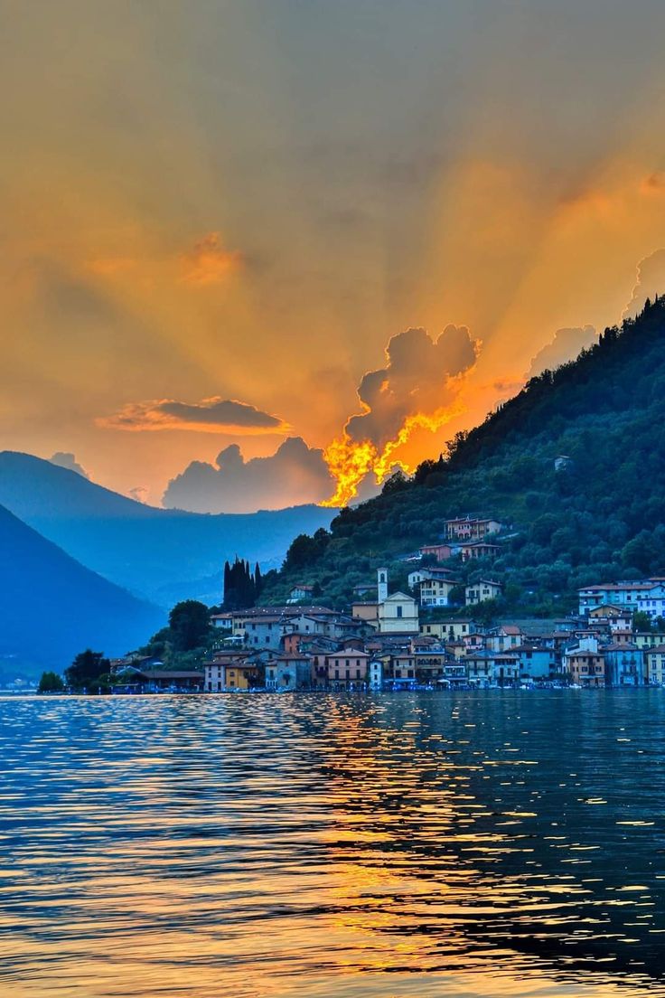 the sun is setting over a small town by the water's edge with mountains in the background