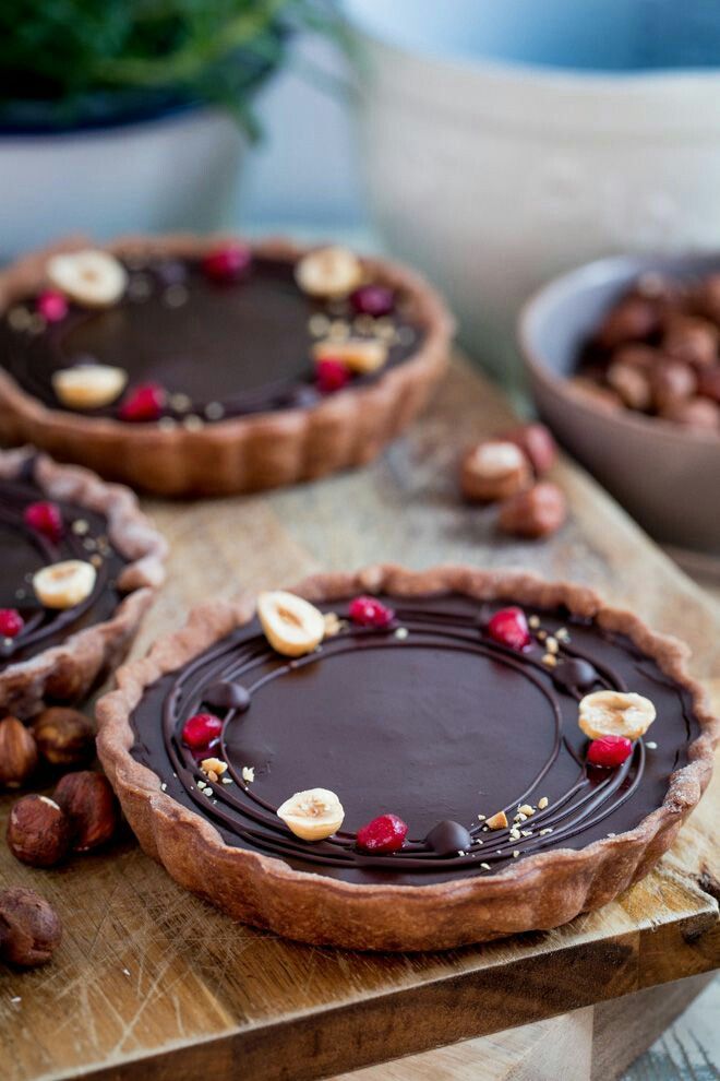 three chocolate tarts with nuts and cherries on a cutting board next to bowls