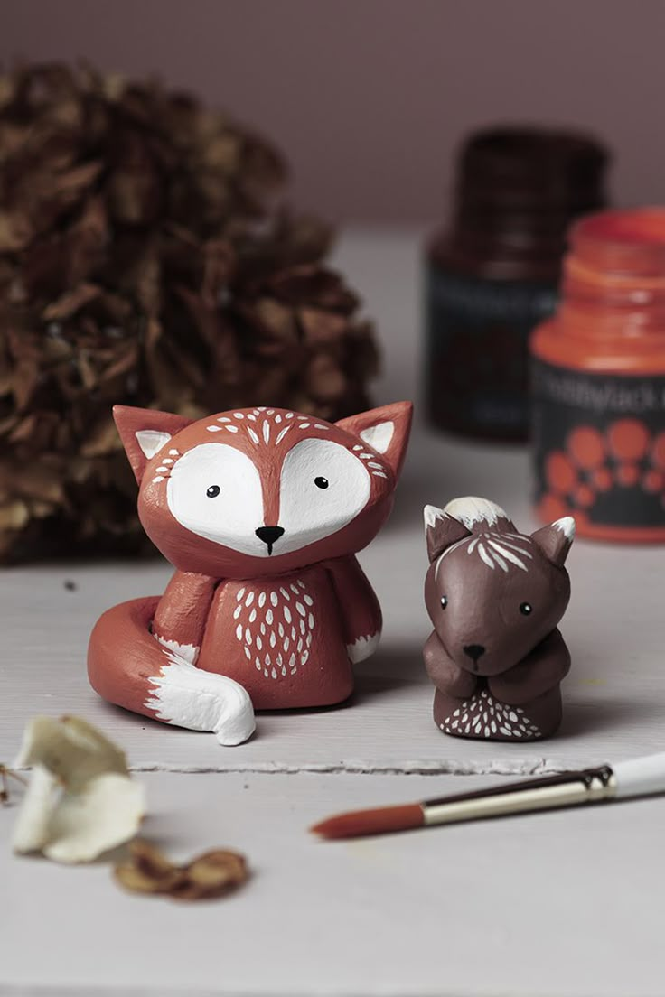 two clay animals sitting on top of a table next to paintbrushes and pine cones