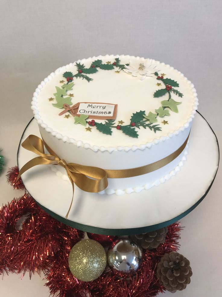 a white cake decorated with holly and pine cones on top of a platter surrounded by christmas decorations