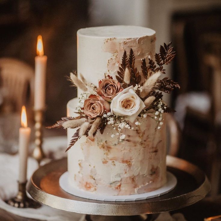 a white wedding cake with flowers and feathers on top sitting on a table next to two candles