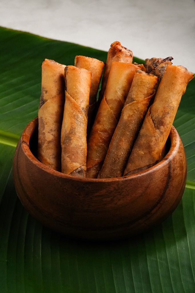 a wooden bowl filled with food on top of a leafy green tablecloth next to a banana leaf