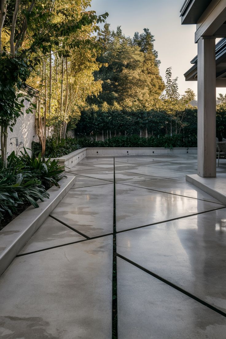 Modern concrete patio with geometric design, surrounded by greenery and shaded by nearby trees. Patio With Concrete Squares, Concrete That Looks Like Pavers, Patio Hardscape Ideas Design, Concrete Courtyard Ideas, Concrete With Grass In Between, Creative Concrete Ideas, Small Concrete Backyard Ideas, Concrete Backyard Ideas Cement Patio, Modern Concrete Patio Ideas